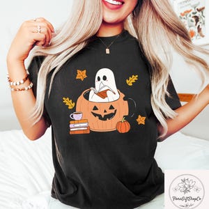 May include: Black t-shirt featuring a Halloween-themed graphic. The design includes a ghost reading a book inside a carved pumpkin, surrounded by autumn leaves and a small pumpkin. A stack of books and a cup of coffee are also included.