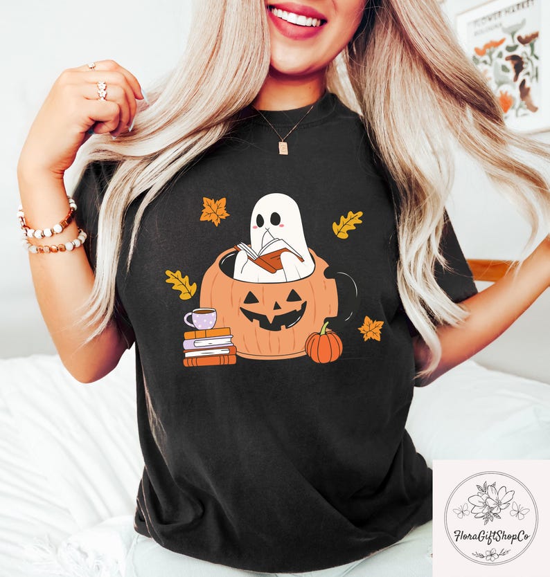 May include: Black t-shirt featuring a Halloween-themed graphic. The design includes a ghost reading a book inside a carved pumpkin, surrounded by autumn leaves and a small pumpkin. A stack of books and a cup of coffee are also included.