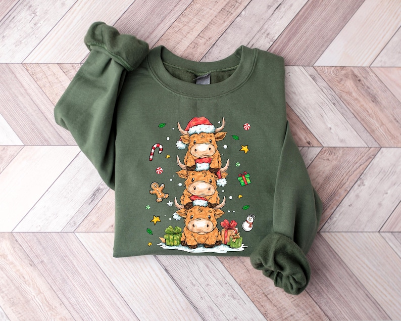May include: An olive green sweatshirt with a festive design of three Highland cows wearing Santa hats, stacked on top of each other. The design includes Christmas decorations like a candy cane and wrapped gifts. The sweatshirt has long sleeves.