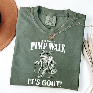 May include: An olive green t-shirt with the text "IT'S NOT A PIMP WALK IT'S GOUT!" and a cartoon frog wearing a hat, coat, and holding a cane. A gold necklace is draped across the shirt. A brown hat and dried plant are in the background.