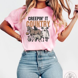 May include: Pink t-shirt with the text "CREEPIN' IT COUNTRY" in brown and tan. The graphic features a horse, cat, cow, pig, and chicken, all in Halloween costumes. The shirt is made of a soft material.