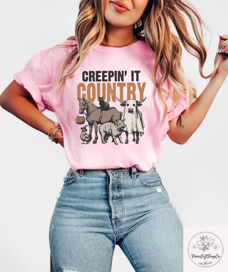 May include: Pink t-shirt with the text "CREEPIN' IT COUNTRY" in brown and tan. The graphic features a horse, cat, cow, pig, and chicken, all in Halloween costumes. The shirt is made of a soft material.