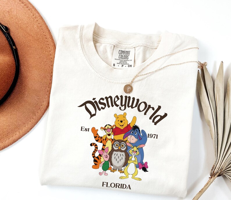 Pode incluir: Uma t-shirt de manga curta cor de creme com um gr&aacute;fico do Winnie the Pooh e amigos. O texto diz Disneyworld Est 1971 FLORIDA. A t-shirt tem uma etiqueta da marca Comfort Colors no pesco&ccedil;o e &eacute; tamanho S. O gr&aacute;fico inclui o Tigre, o Piglet, o Pooh, o Coruja, o Igor e o Coelho.