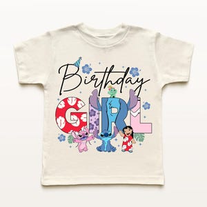 Op de afbeelding: Cr&egrave;me-kleurig T-shirt met korte mouwen met de woorden "Birthday Girl" in zwart script. Het woord "GIRL" staat in kleurrijke blokletters met stripfiguren. De shirt heeft een verjaardagshoed en bloemmotieven.