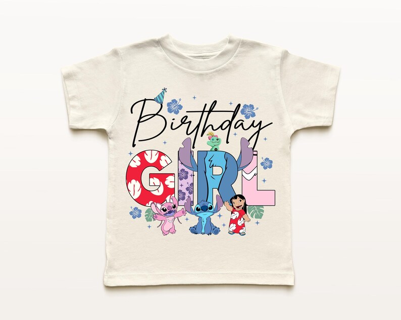 Op de afbeelding: Cr&egrave;me-kleurig T-shirt met korte mouwen met de woorden "Birthday Girl" in zwart script. Het woord "GIRL" staat in kleurrijke blokletters met stripfiguren. De shirt heeft een verjaardagshoed en bloemmotieven.