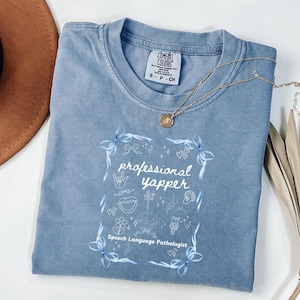 Puede incluir: Una camiseta azul Comfort Colors con las palabras "professional yapper" y "Speech Language Pathologist" impresas en la parte delantera. La camiseta tiene cuello redondo y mangas cortas. Un collar dorado con un colgante está encima de la camiseta.
