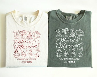 Può includere: Due magliette piegate, una color crema e una grigio-verde, con una grafica a tema natalizio. La maglietta color crema ha un design rosso, e la maglietta grigio-verde ha un design bianco. Entrambe le magliette dicono "Merry & Married" con nome personalizzato e "EST 2025".