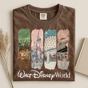 May include: Brown t-shirt with a graphic of Walt Disney World landmarks. The design includes images of a tree, a castle, a geodesic sphere, and a hotel. The words "Walt Disney World" are printed in a cursive font at the bottom of the shirt.