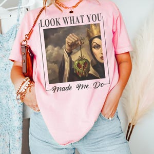 May include: A light pink short-sleeve t-shirt featuring a graphic print of a queen character holding a dripping green apple. The text above the image reads LOOK WHAT YOU and the text below the image reads Made Me Do in a black serif font.