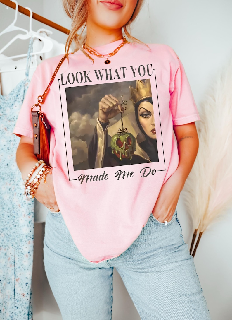 May include: A light pink short-sleeve t-shirt featuring a graphic print of a queen character holding a dripping green apple. The text above the image reads LOOK WHAT YOU and the text below the image reads Made Me Do in a black serif font.