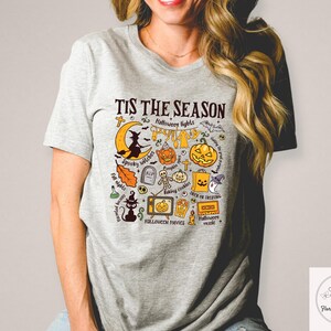 Pu&ograve; includere: T-shirt grigio chiaro con grafica a tema Halloween. Il design include una luna crescente con una strega, zucche, pipistrelli e la scritta "Tis the Season". Altri elementi includono un gatto nero, una lapide e una televisione.