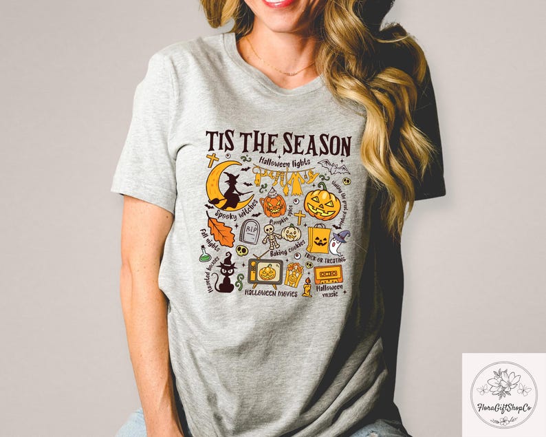 Pu&ograve; includere: T-shirt grigio chiaro con grafica a tema Halloween. Il design include una luna crescente con una strega, zucche, pipistrelli e la scritta "Tis the Season". Altri elementi includono un gatto nero, una lapide e una televisione.