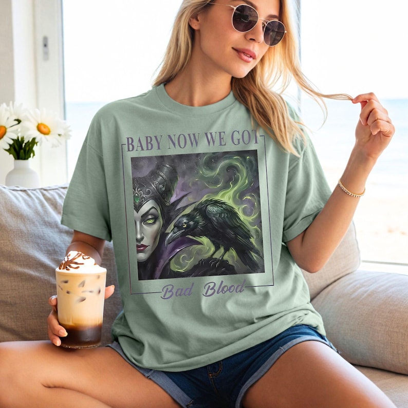 May include: A sage green t-shirt featuring a graphic print of a dark-haired character with horns and a black raven surrounded by green smoke. The text "BABY NOW WE GOT" is printed above the image, and "Bad Blood" is printed below the image.