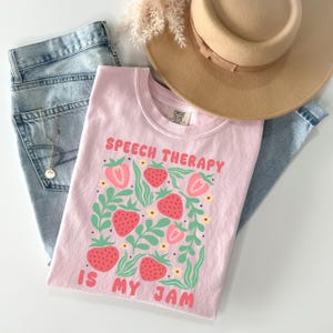 May include: A light pink t-shirt with the text "SPEECH THERAPY IS MY JAM" and a strawberry design. The shirt is folded on top of blue jeans, a tan hat, and a necklace.