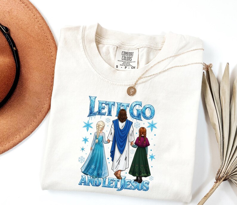 May include: Cream-colored t-shirt with the text "Let It Go and Let Jesus" in blue. The graphic features Jesus holding hands with two cartoon characters. A gold necklace and a brown hat are also visible.