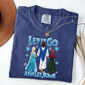 May include: A navy blue t-shirt with the text "LET IT GO AND LET JESUS" in blue and white. The graphic depicts Jesus with two girls in blue and green dresses. A gold necklace and a brown hat are also visible.