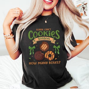 Puede incluir: Camiseta negra con el texto "Sorry, Can't. Cookies Season" en verde y naranja. La camiseta presenta ilustraciones de tres galletas diferentes y lazos verdes. La frase "How Many Boxes?" est&aacute; impresa debajo.