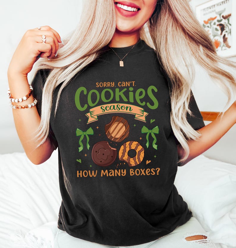 Puede incluir: Camiseta negra con el texto "Sorry, Can't. Cookies Season" en verde y naranja. La camiseta presenta ilustraciones de tres galletas diferentes y lazos verdes. La frase "How Many Boxes?" est&aacute; impresa debajo.