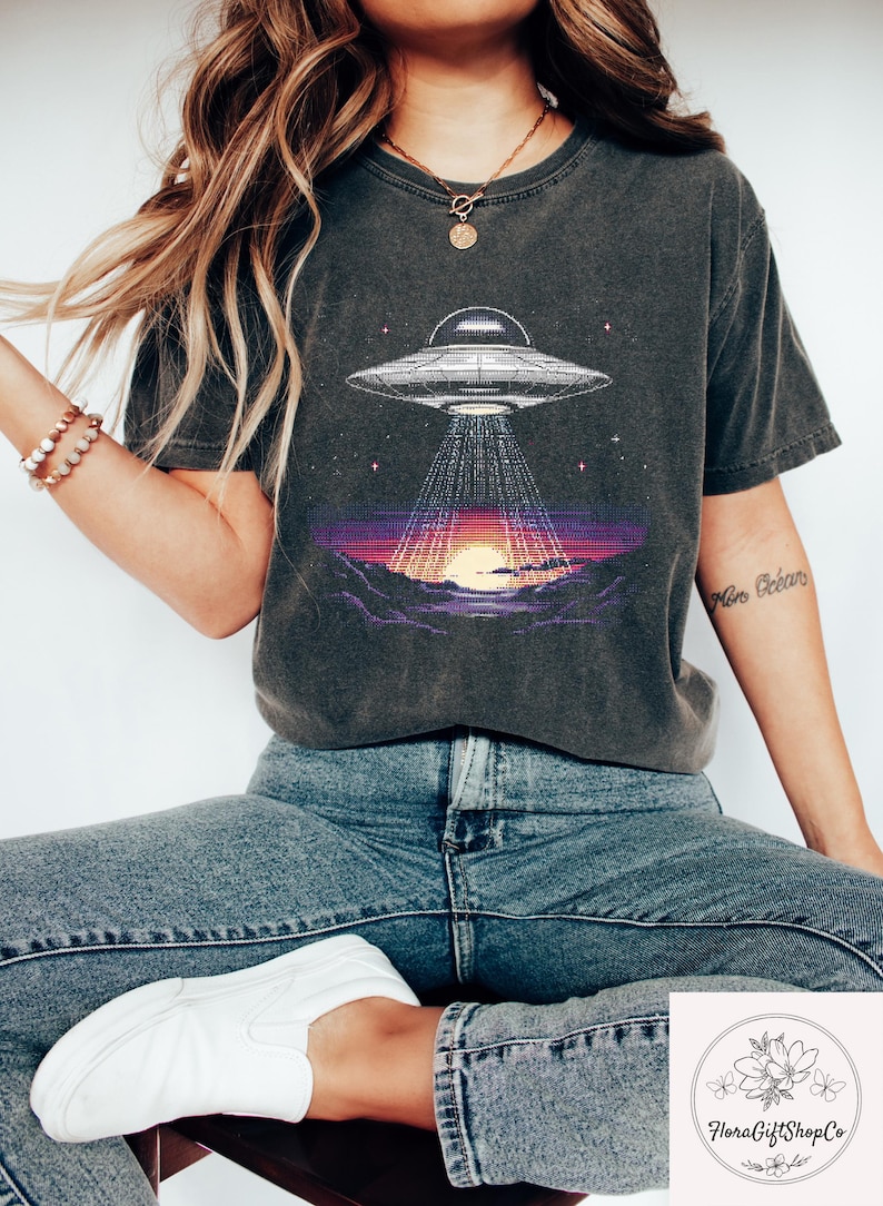 Op de afbeelding: Donkergrijs T-shirt met een gepixeld grafisch ontwerp van een UFO die licht schijnt op een zonsondergang. Het ontwerp omvat een gedetailleerd ruimteschip en een levendige zonsondergang met paarse en oranje tinten. Het is een casual kledingstuk.