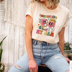 May include: Cream-colored t-shirt with the text "Counseling is My Jam" in a retro font. The graphic features cartoon characters of a slice of bread and a jar of jam. The shirt is being worn by a person.