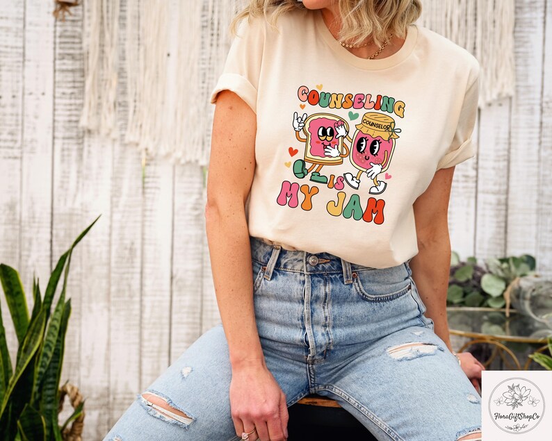 May include: Cream-colored t-shirt with the text "Counseling is My Jam" in a retro font. The graphic features cartoon characters of a slice of bread and a jar of jam. The shirt is being worn by a person.