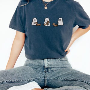 May include: A navy blue t-shirt featuring an embroidered design of three cartoon ghosts with dachshund dogs. The ghosts are in different poses, holding or walking the dogs. The shirt is a casual style.
