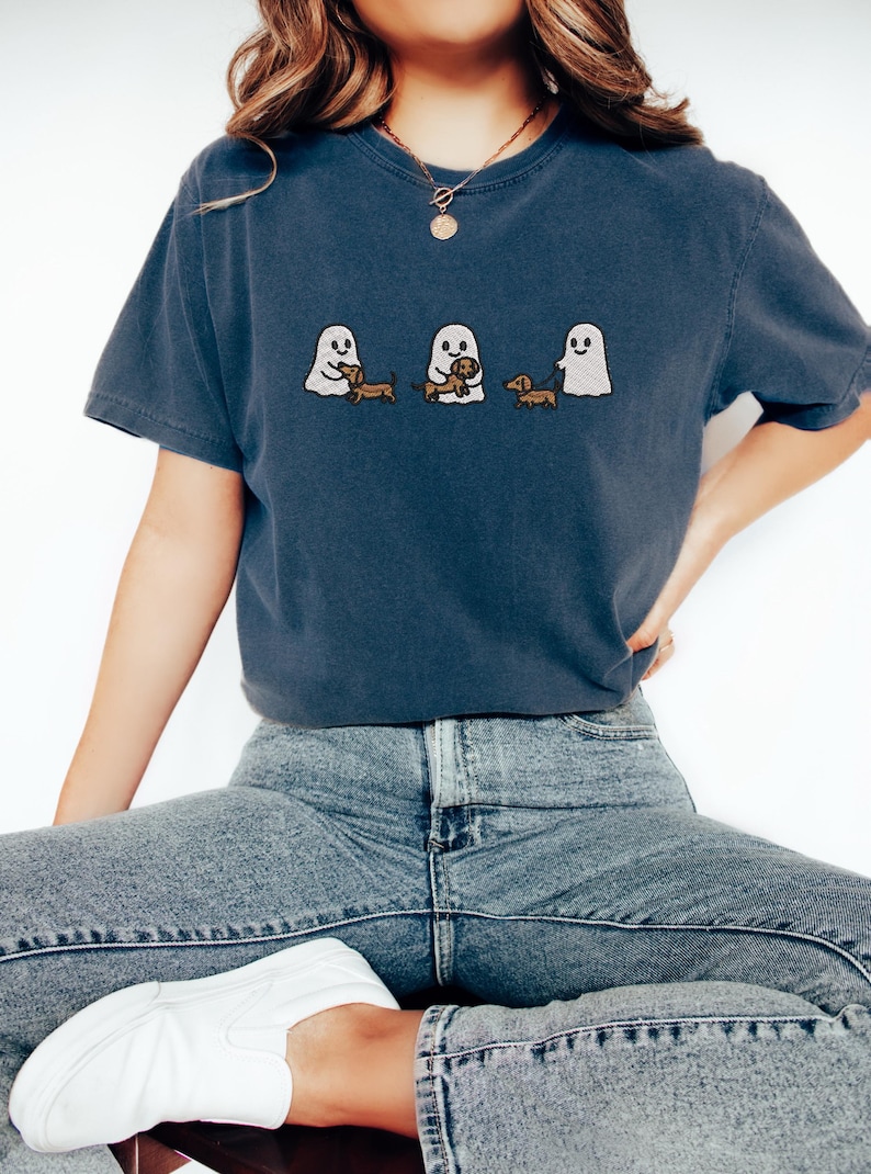 May include: A navy blue t-shirt featuring an embroidered design of three cartoon ghosts with dachshund dogs. The ghosts are in different poses, holding or walking the dogs. The shirt is a casual style.