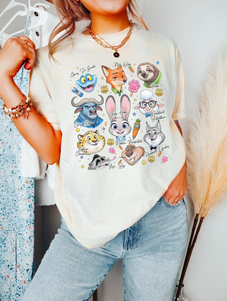 Puede incluir: Una camiseta de manga corta color crema con un estampado gr&aacute;fico colorido de varios personajes animales animados de Zootopia, incluidos Judy Hopps, Nick Wilde y Flash, junto con peque&ntilde;os iconos como zanahorias, huellas y placas policiales.