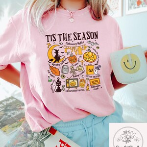 Pu&ograve; includere: T-shirt rosa con grafica a tema Halloween e la scritta "TIS THE SEASON". Il design include una strega, zucche e le parole "Spooky witches" e "Trick or Treating".