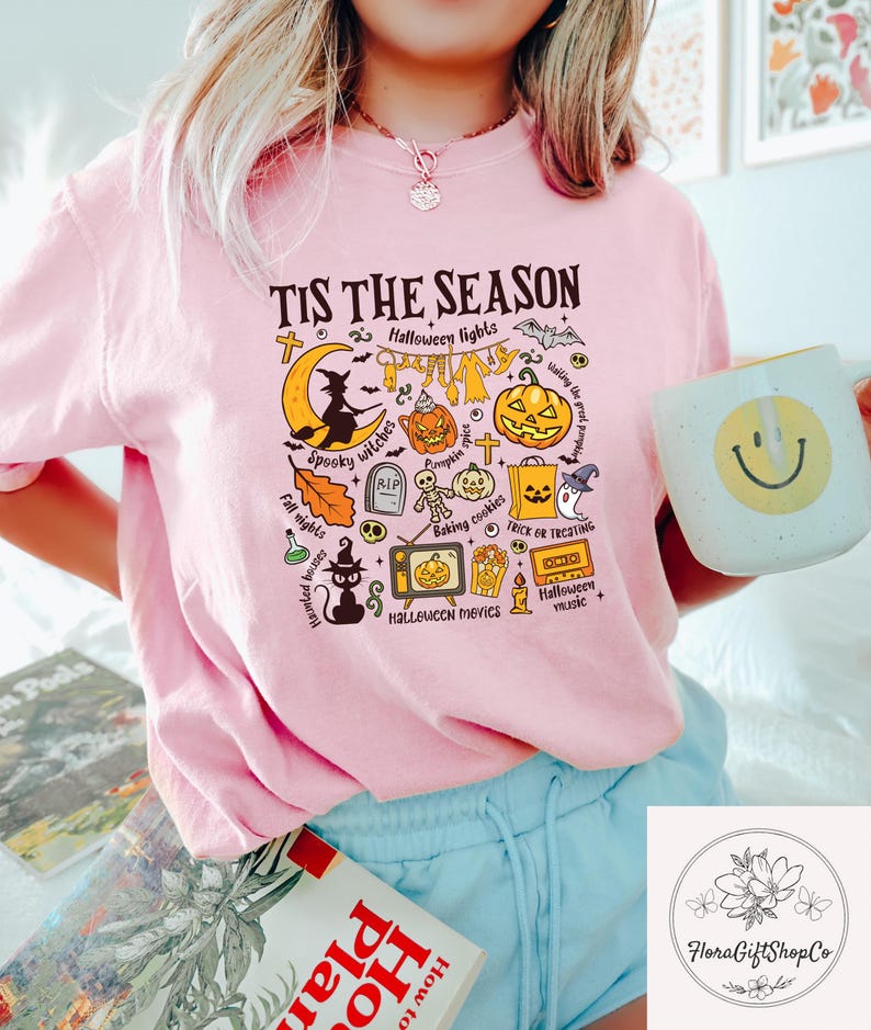Pu&ograve; includere: T-shirt rosa con grafica a tema Halloween e la scritta "TIS THE SEASON". Il design include una strega, zucche e le parole "Spooky witches" e "Trick or Treating".
