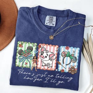 May include: A navy blue t-shirt featuring a graphic print of Moana, Pua the pig, and various themed icons within three rectangular frames. The text "There's just no telling how far I'll go" is printed in white script below the colorful illustrations.