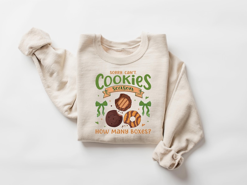 Puede incluir: Sudadera color crema con el texto "Sorry, Can't Cookies Season" en verde y naranja. El dise&ntilde;o incluye ilustraciones de tres galletas y lazos verdes. La frase "How Many Boxes?" est&aacute; debajo.