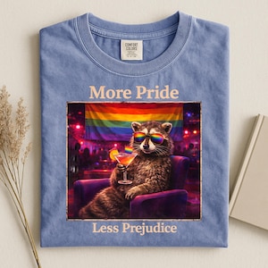 Op de afbeelding: Lichtblauw t-shirt met de woorden "More Pride" en "Less Prejudice" gedrukt boven en onder een ingelijste afbeelding van een wasbeer met een regenboog zonnebril en een cocktail. De afbeelding bevat een regenboogvlag.