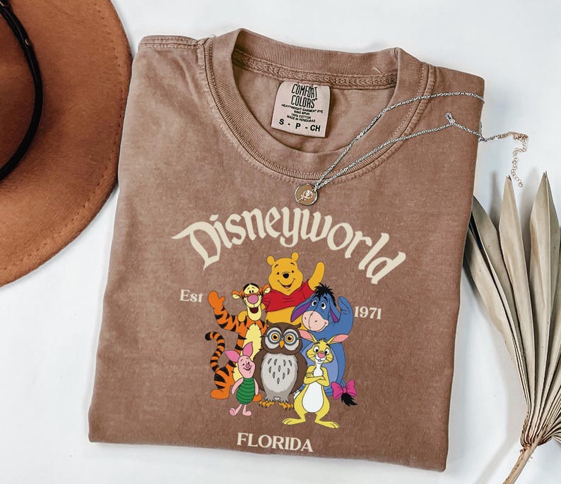 Pode incluir: Uma t-shirt castanha de manga curta com um gr&aacute;fico do Winnie the Pooh, Tigre, Piglet, Eeyore, Owl e Rabbit. O texto diz Disneyworld Est 1971 Florida. A t-shirt tem uma etiqueta da marca Comfort Colors na parte interna da gola.