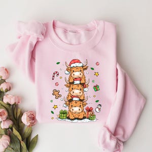 May include: A pale pink sweatshirt with a festive design. It features a stack of three cartoon Highland cows, each wearing a Santa hat and scarf. Christmas decorations like a candy cane and gifts surround the cows.