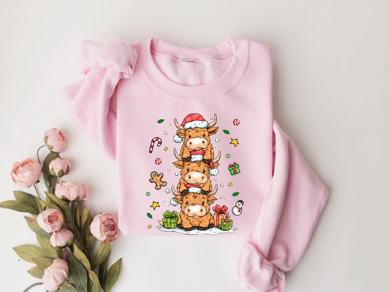 May include: A pale pink sweatshirt with a festive design. It features a stack of three cartoon Highland cows, each wearing a Santa hat and scarf. Christmas decorations like a candy cane and gifts surround the cows.