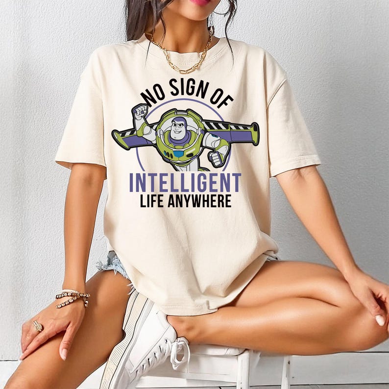 May include: Beige t-shirt featuring a graphic of Buzz Lightyear with the text "NO SIGN OF INTELLIGENT LIFE ANYWHERE". The design includes green, purple, and white accents. The shirt has short sleeves and a relaxed fit.