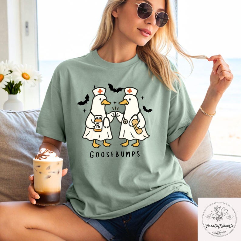 May include: Sage green t-shirt featuring a cartoon design of two ducks dressed as nurses, with bats and stars. The ducks are fist-bumping, and the text "GOOSEBUMPS" is below the graphic. The shirt is a relaxed fit.