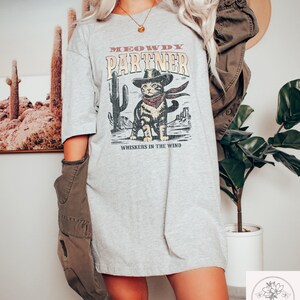 K&ouml;nnte beinhalten: Hellgraues T-Shirt mit einem Aufdruck einer Katze mit Cowboyhut und Halstuch, mit dem Text "MEOWDY PARTNER" und "WHISKERS IN THE WIND". Das Design umfasst Kakteen und eine W&uuml;stenlandschaft.
