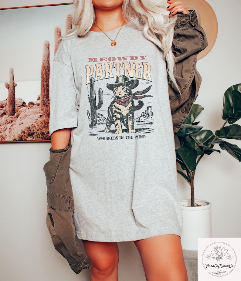 K&ouml;nnte beinhalten: Hellgraues T-Shirt mit einem Aufdruck einer Katze mit Cowboyhut und Halstuch, mit dem Text "MEOWDY PARTNER" und "WHISKERS IN THE WIND". Das Design umfasst Kakteen und eine W&uuml;stenlandschaft.