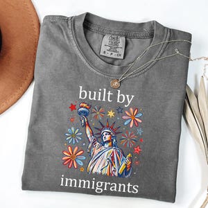 May include: A charcoal gray short-sleeve t-shirt featuring a graphic of the Statue of Liberty surrounded by colorful flowers and star shapes. The text "built by immigrants" is printed in white above and below the central graphic illustration.