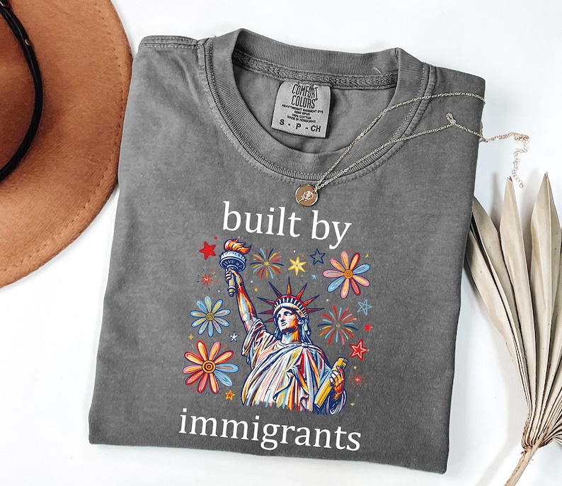 May include: A charcoal gray short-sleeve t-shirt featuring a graphic of the Statue of Liberty surrounded by colorful flowers and star shapes. The text "built by immigrants" is printed in white above and below the central graphic illustration.