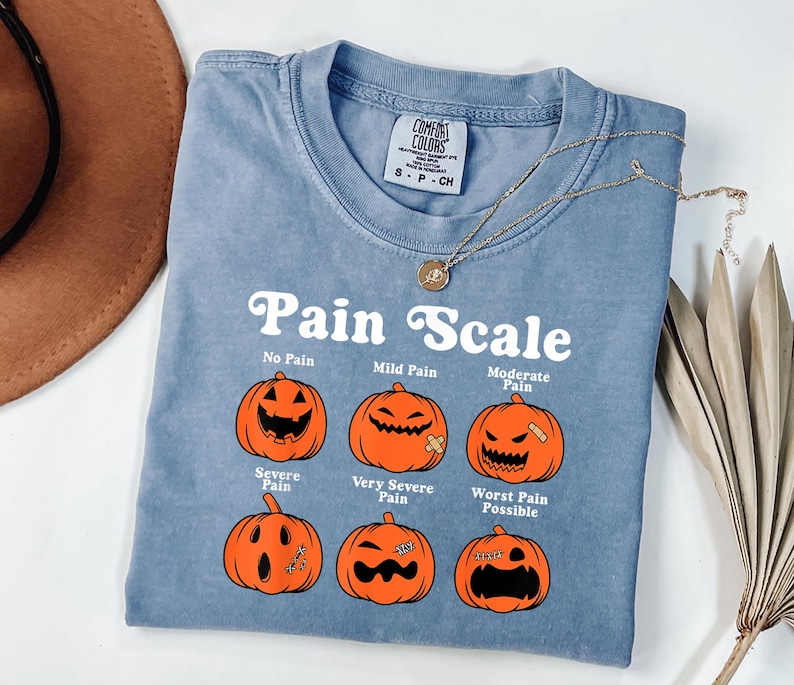 May include: A blue Comfort Colors t-shirt with a "Pain Scale" graphic featuring orange jack-o'-lanterns. The shirt has a crew neck and short sleeves. A gold necklace with a pendant is draped across the shirt.