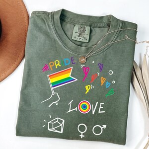 May include: An olive green short-sleeve t-shirt featuring a graphic print with the text #PRIDE and LOVE, a rainbow flag, hand-drawn hearts, a geometric shape, and gender symbols. The shirt has a Comfort Colors brand label at the inner neckline.
