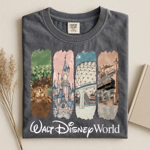 May include: Dark gray t-shirt featuring a colorful graphic of Walt Disney World landmarks. The design includes images of a castle, a tree, a geodesic sphere, and a hotel. The words "Walt Disney World" are printed in white script across the bottom.