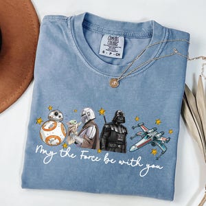 Puede incluir: Camiseta azul claro con personajes de Star Wars: BB-8, The Mandalorian, Darth Vader y un X-wing. La frase "May the Force be with you" está escrita debajo. La camiseta tiene cuello redondo y mangas cortas.