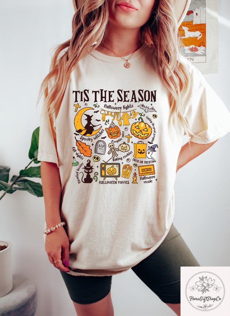 Pu&ograve; includere: T-shirt color crema con grafica a tema Halloween. Il design include una luna crescente, una strega, zucche e la scritta "TIS THE SEASON". Altre illustrazioni includono una lapide, un sacchetto di caramelle e una TV.