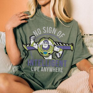 May include: Olive green t-shirt featuring a graphic of Buzz Lightyear with the text "NO SIGN OF INTELLIGENT LIFE ANYWHERE". The design includes Buzz in his spacesuit with purple and green accents. The shirt is a relaxed fit.