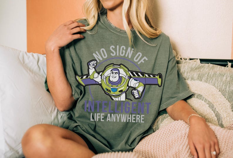 May include: Olive green t-shirt featuring a graphic of Buzz Lightyear with the text "NO SIGN OF INTELLIGENT LIFE ANYWHERE". The design includes Buzz in his spacesuit with purple and green accents. The shirt is a relaxed fit.