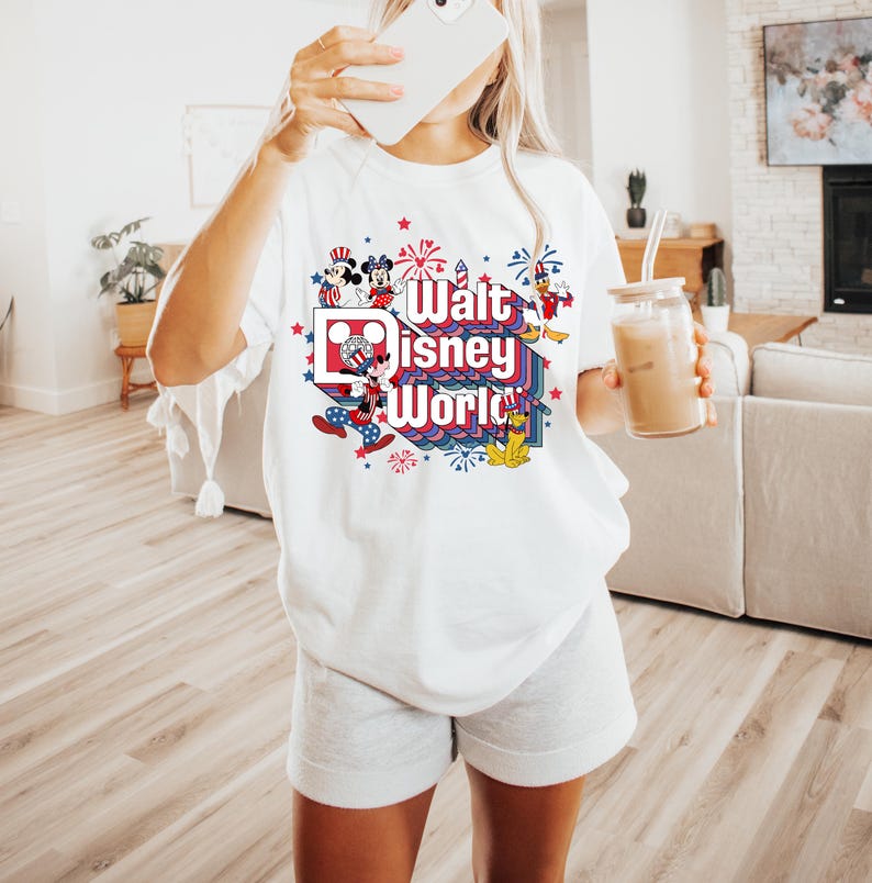 K&ouml;nnte beinhalten: Wei&szlig;es T-Shirt mit einer bunten Grafik mit Mickey und Minnie Mouse, Feuerwerk und dem Text "Walt Disney World". Das Design enth&auml;lt rote, wei&szlig;e und blaue Elemente. Das Shirt ist ein l&auml;ssiger Stil.